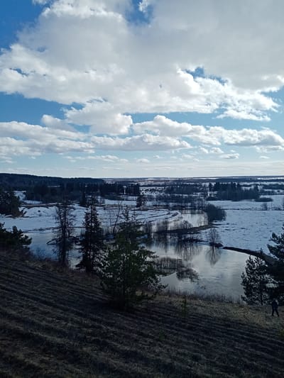 The image depicts a landscape in Сундур, Russia, likely during springtime given the melting snow. The scene is viewed from an elevated position, looking down on a body of water reflecting the sky. Patches of snow are scattered across the ground and among trees. There's a forest in the mid-ground. The sky is predominantly blue, covered in cumulus clouds. On a grassy hillside in the lower-right corner, a person is walking.