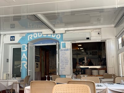 The image shows the exterior of a restaurant in Tamariu, Spain. The restaurant is called "Rodondo Bar". It has a blue and white exterior with a sign that reads "Rodondo Bar" in blue lettering on a white background. The sign is located above a door with a window. The window is covered with a white curtain. The door is open and there is a table and chairs inside. There are two security cameras visible on the exterior of the restaurant. The restaurant is located on a street. There are several tables set up outside of the restaurant with chairs. Some of the tables are covered with white tablecloths, while others are not. There are glasses on the table, but no food. The restaurant is located at 41.918, 3.208 in Tamariu, Spain. The tables are arranged in an informal manner. There is a sign on the wall next to the door, advertising the restaurant's menu. There are people inside the restaurant, seemingly waiting on tables. The restaurant is surrounded by a white awning and a light-colored tiled floor. The image appears to have been captured on a sunny day, as shadows are cast on the floor.