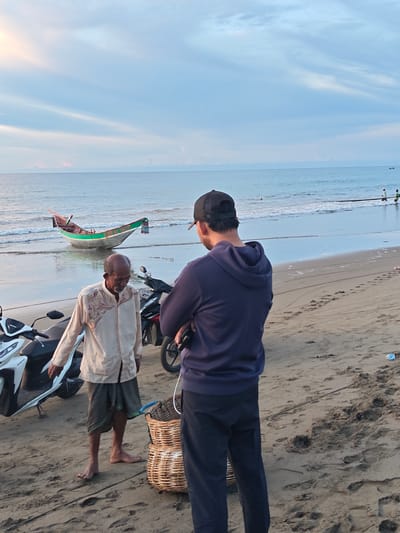 Two men stand on a sandy beach in Uleekareung, Indonesia, near the ocean. The man on the left, appearing to be older, wears a cream-colored, long-sleeved shirt with ornate embroidery and a dark, checkered sarong. He is barefoot. To his right, a younger man wearing a dark blue hooded sweatshirt and a black baseball cap faces away from the camera, with his arms crossed and holding a phone in his right hand. In front of the older man are two stacked woven baskets. Several motorcycles are parked on the sand behind the men. In the background, a small fishing boat with green and red accents rests on the wet sand at the edge of the water. The ocean stretches to the horizon under a pale blue sky with wispy clouds. Two other figures are visible in the distance in the water, possibly engaged in fishing activities. The overall scene suggests a coastal, possibly early morning or late afternoon, setting.