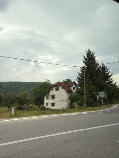 The image depicts an exterior view of a rural scene in Rastovci, Bosnia and Herzegovina, taken from a moving vehicle. The primary subjects are an old, partially derelict two-story building with a red tile roof, surrounded by dense green foliage and trees. A paved road with a white lane marking runs alongside the property, indicating the perspective of a roadside observer or passenger.
The building appears weathered, with some visible damage and blank window frames. The surrounding vegetation is lush, suggesting a natural, possibly overgrown, environment. In the background, a heavily forested hillside rises, creating a sense of depth and the typical landscape of the region. The sky is overcast with visible clouds, implying a daytime scene with potentially cloudy or impending rainy weather.
A wooden pole with power lines is visible to the left of the frame, adding to the rural setting. A road sign with a circular shape and a red bar is also present, positioned behind some trees near the road. The overall atmosphere is one of quietness and perhaps a touch of melancholy due to the state of the building and the subdued lighting. No people are visible in the image, and no text is discernibly legible. The image is oriented vertically, with the road and building running along the right side.