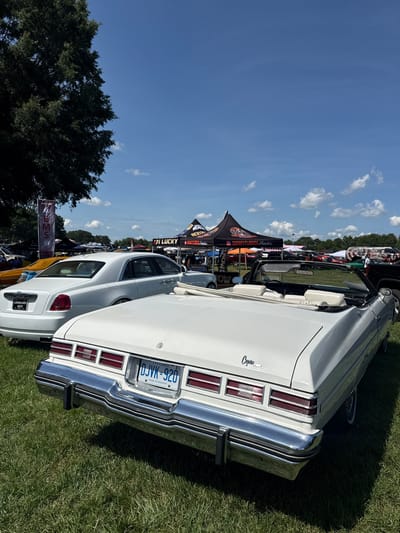The image shows a white convertible parked on a grassy field at the Georgia International Horse Park in Fairburn, United States, under a clear blue sky with scattered clouds. In the foreground, the rear of the white convertible is visible, featuring a chrome bumper, red taillights, and a license plate that reads "DJVK 920 ONTARIO". The word "Caprice" is visible on the trunk. To the left, the rear of a white sedan, possibly a Rolls-Royce, is also visible. In the background, several tents and other vehicles are present, suggesting an outdoor event or car show. A large tree with dark green foliage is on the left side of the image.