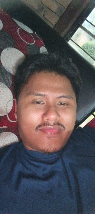 A close-up, slightly angled selfie of a young man is captured from the chest up. He has dark hair, brown eyes, and a mustache and light stubble. He is wearing a dark blue shirt. He is positioned against a background that includes a patterned fabric with circles in red, gray, and white, and a dark surface that could be furniture. To the right, a window with a view of greenery and a brick wall is visible. The lighting suggests an indoor setting, possibly during the daytime. The location in Kemuning, Indonesia is not discernible from the image itself.