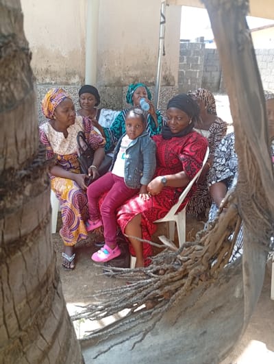 An outdoor scene in Unguwar Gaya, Nigeria, depicts eight individuals seated, comprising seven adult females and one young female child. All are seated on white plastic chairs, positioned under a partial white canopy structure visible via a support pole.
In the center, a young female child, approximately 3-5 years old, is seated on the lap of an adult female. The child wears a white t-shirt, a blue denim jacket, red trousers, and pink clogs, and looks directly towards the viewer. The adult female holding the child wears a red and black patterned dress, a black headwrap, and a wristwatch on her left wrist; she is looking downward and slightly to her right. To this adult's immediate left, another adult female wears a yellow, purple, and white patterned dress with a matching headwrap, and looks to her own left. A black handbag rests on her lap. Directly behind the child, a third adult female, dressed in a green and white patterned outfit and a green headwrap, is drinking from a clear plastic water bottle. Four additional adult females, similarly clad in various traditional patterned garments and headwraps, are seated around the periphery of the group.
The background consists of a weathered, light-colored concrete or plastered wall, transitioning to a grey cinder block wall further to the right. The ground beneath the individuals is exposed earth. The left foreground is partially obscured by a large, vertically textured tree trunk, possibly a palm tree. In the lower right foreground, a cluster of dried, fibrous palm frond bases is visible. Strong sunlight indicates a daytime setting.