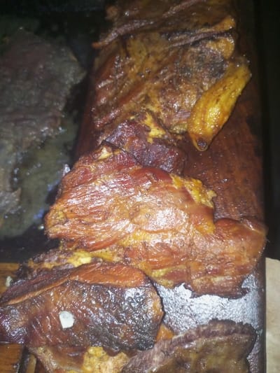 Assorted cuts of cooked meat are arranged on a wooden board. Some pieces appear more charred, while others have a lighter, golden-brown color. The meat texture suggests it might be brisket or a similar slow-cooked cut. There are visible fat marbling and caramelized surfaces, indicating rich flavor and juiciness. Some pieces are glistening, likely from rendered fat. A dark area beside the wooden board might indicate additional meat or a side dish.