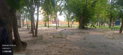 A daytime outdoor shot shows a dirt and gravel area with scattered trees and vegetation. In the background, a light-colored building with an ornate yellow trim and a red-tiled roof is visible. The area appears to be part of a temple or religious complex. The ground is uneven and covered with sand, small stones, and sparse green plants. The foliage is dense in some areas, creating shade. A dark structure, possibly a building, is visible on the far right. The timestamp "2026/06/18 09:18" and the brand logo "TECNO" are overlaid on the bottom left of the image.