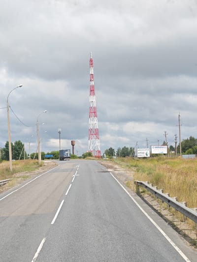 The image captures a roadside scene in the city of Saltykovo, Russia. A tall, red and white radio tower dominates the skyline, with a water tower visible in the middle ground. The road, marked with white lines, curves to the right, bordered by dry grass and vegetation on either side. A grey truck is driving away from the viewer on the road. Several streetlights line the left side of the road. In the distance, trees form a dense line along the horizon. Billboards are visible on the right side of the road, one with text in Cyrillic script. The sky is overcast with grey clouds.