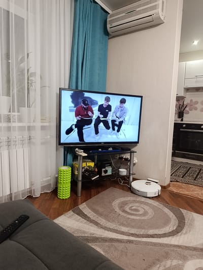 The image depicts an indoor living room scene in Ishim, Russia. A large television screen displays three young men, two of whom appear to be seated while one is standing. The room is furnished with a dark grey sofa in the foreground, a glass-top media console with various electronic devices, and a vibrant green foam roller. White sheer curtains and teal drapes are visible in the background. An air conditioning unit is mounted high on the wall. A white robot vacuum cleaner is positioned on the floor near the media console. The lighting suggests it is daytime. The overall atmosphere appears relaxed and domestic, with a focus on entertainment and comfort.