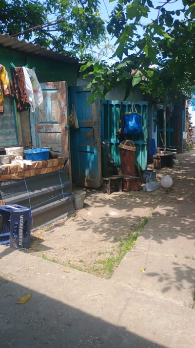 The image depicts a rustic, outdoor scene in what appears to be a yard or alleyway in Mitoc, Moldova. A weathered, blue-painted structure with a corrugated metal roof runs along the back of the scene. Clothes hang on a line in front of one of the weathered wooden doors. Various objects are scattered around, including a blue plastic crate labeled "FLENSBURGER" in the lower left corner, buckets, large plastic water jugs, and a reddish-brown cylindrical object. Wooden stools and benches are placed near the structure. The ground is a mixture of dirt and concrete, with patches of grass. Sunlight casts dappled shadows from the overhanging trees, suggesting a bright, possibly warm day. The overall impression is one of a humble, lived-in space.