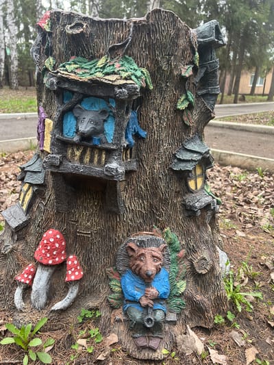 The image shows a decorative tree stump, potentially located in Sanatoriya Yumatovo Imeni 15-Letiya Bassr, Russia (as per provided location). The stump features various molded and painted elements, including a sleeping figure resembling a hedgehog or similar creature wearing a blue shirt, a mouse-like figure within a dwelling, red-capped mushrooms, and a small window. The environment surrounding the stump consists of dirt, leaves, and some vegetation, with a paved path and building visible in the blurred background. The photograph is taken at a relatively close range, providing a detailed view of the stump's textured surface and painted components.