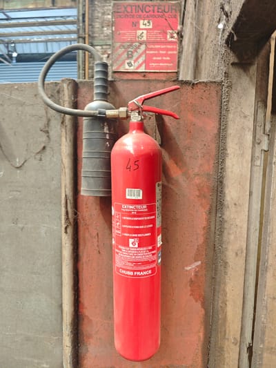 The image shows a red CO2 fire extinguisher mounted on a dirty, reddish-orange wall in what appears to be an industrial or workshop setting. The extinguisher has a black hose with a black nozzle attached. A small red label on the extinguisher reads "EXTINCTEUR 5 KG DIOXYDE DE CARBONE". The number "45" is written in black on the body of the extinguisher. A white barcode is also visible on the lower part of the extinguisher. A red sign is mounted on the wall above the extinguisher, with French text reading "EXTINCTEUR DIOXYDE DE CARBONE N° 45" and "CLASSE B". The environment appears weathered and dusty, with cobwebs visible in the upper right portion of the image. The lighting is somewhat dim, suggesting an indoor location. No people are visible in the image.
