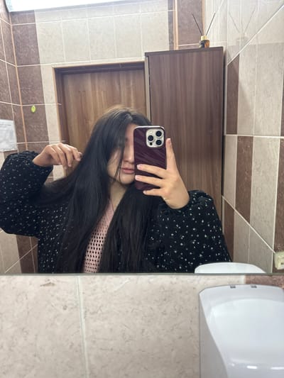 A young woman with long, dark hair is taking a selfie in a bathroom. She's holding a smartphone, partially obscuring her face. She's wearing a black dress with small white floral patterns and a pink undershirt. The bathroom has brown and beige tiled walls, a wooden cabinet, and a white hand dryer. The scene is dimly lit. Location: Davydovo, Russia.