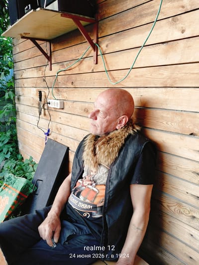 An outdoor shot taken in Oseyevo, Russia, shows a bald, middle-aged Caucasian man with weathered skin sitting on what appears to be a bench or ledge against a wooden wall. He is wearing a black vest with a fluffy fur collar over a t-shirt with a graphic print. The man is looking to his left, with his head tilted slightly upwards, and his expression is neutral. A tattoo is visible on his left forearm.

Behind him, the wooden wall is constructed from horizontally laid planks. A white electrical outlet with three sockets is mounted on the wall, with a black and a green cord plugged in. A shelf with electronic equipment is attached to the wall above the outlet. To the left of the man, lush green foliage is growing, partially obscuring a green and white bag.

The lighting suggests it is daytime, with soft shadows cast on the wooden wall, indicating a possibly overcast or late afternoon light. The overall scene evokes a relaxed, casual outdoor setting. The bottom right corner of the image displays text that reads "realme 12 24 июня 2026 г. в 19:24".