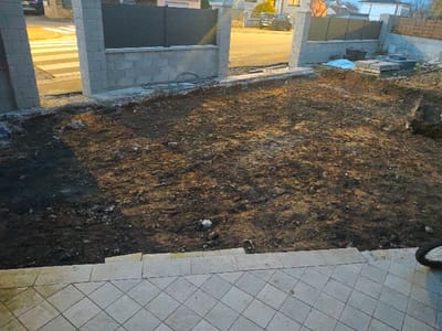 The scene depicts an outdoor residential property under construction or renovation in Aumetz, France, observed from an elevated perspective. In the immediate foreground, a light grey tiled surface, composed of square pavers in a grid pattern, leads to two shallow steps descending into a disturbed plot of ground. The central midground consists of an uneven expanse of dark brown earth, interspersed with lighter soil, scattered small rocks, and debris, indicative of recent excavation or landscaping.

The background features a perimeter fence, partially constructed with two vertical grey concrete block pillars. These pillars flank a lower grey concrete block wall, which is topped with dark grey opaque fence panels. Beyond this fence, a public asphalt road is visible. On the left side of the road, a white pedestrian crossing is present, and a dark-colored vehicle is partially visible. To the right of the fence, within the property boundaries, stacks of grey paving blocks are present, and a blue tarp covers additional construction materials. Residential structures with pitched roofs are discernible in the distant background. The environment is illuminated by a combination of natural ambient light, consistent with twilight or dawn, and artificial lighting from street sources, casting varied light and shadow patterns. No individuals are present in the image. A bicycle tire is partially visible on the far right edge of the foreground steps.