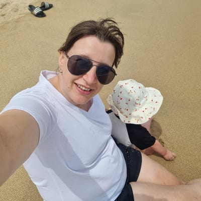 The image shows two people on a sandy beach. The person in the foreground is a woman wearing sunglasses, a white t-shirt, and black shorts. She is smiling and looking at the camera. To her right, a baby is sitting on the sand, wearing a white hat with a cherry pattern and a black and white outfit. In the background, a pair of black slides with white stripes are visible on the sand. The overall environment is a beach with fine sand and the faint presence of water at the edge of the frame, suggesting a coastal location. This scene is consistent with a typical recreational area in Gangneung-si, South Korea.