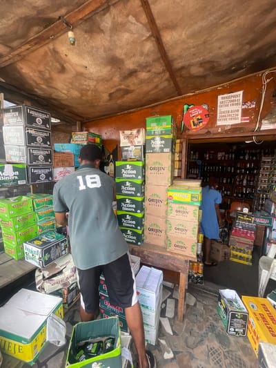 The image depicts the interior of a small shop in Akowonjo, Nigeria, stocked with numerous boxes of beverages. A man wearing a green polo shirt with the number "18" on the back and black shorts is standing in the center, facing away from the camera and interacting with the merchandise. A woman in a blue dress is visible in the background, also within the shop. The shop appears to be an open-air establishment with a wooden ceiling. Stacks of various boxed drinks, including "Action Bitters," "Orijin Bitters," and "Gbefun," are prominently displayed. Some boxes are open, and others are neatly stacked, suggesting inventory or preparation for sale. The ground is paved with a patterned tile. Posters and advertisements are visible on the walls and the shop's exterior.