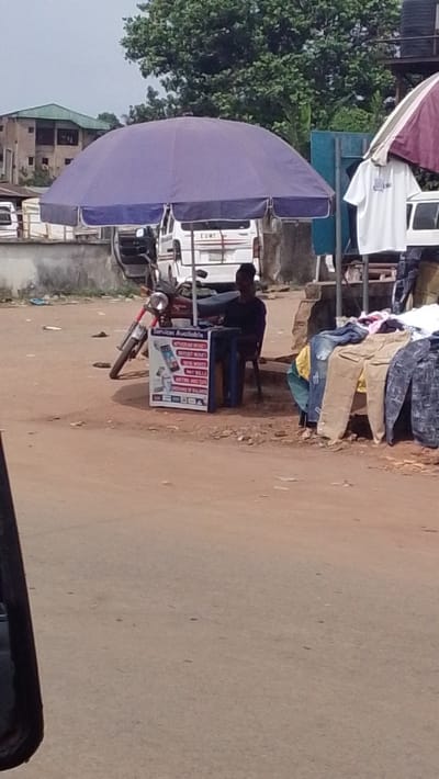 This image depicts a roadside commercial area in Igbo Ukwu, Nigeria, characterized by informal stalls and activity on an unpaved, reddish-brown dirt road under an overcast sky.

In the midground, a large purple market umbrella dominates the left side, providing shade. Beneath it, a person, dressed in a dark top, is seated on a dark chair behind a small dark table. Adjacent to this person is a white rectangular sign with "Services Available" prominently displayed at the top, listing various financial or mobile services, accompanied by illustrations of a smartphone and a Point-of-Sale (POS) terminal. A red motorcycle is parked on its kickstand to the left of the umbrella.

To the right, another market stall displays clothing. A white t-shirt hangs from a frame, and several pairs of trousers or jeans in different colors (including khaki, blue, and patterned) are laid out on a surface. Part of a striped maroon and white umbrella is visible above this stall.

The background reveals a mix of urban elements: multiple white vans or buses are parked or moving, one showing "LUNT" on its rear. Further back, multi-story buildings with green roofs are partially visible through dense green foliage and trees. The foreground contains a dark, out-of-focus object, possibly a vehicle's side panel or mirror, suggesting the photograph was taken from a car.