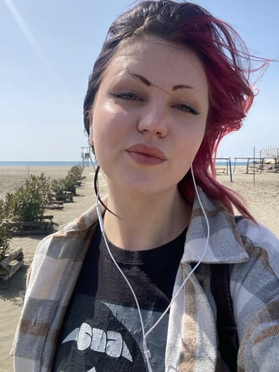 An outdoor image depicts a female individual in the foreground, taking a selfie on a sandy beach under clear, bright blue skies. The subject is a young adult female with light skin, dark hair on her left side, and windblown magenta hair on her right side. She wears dark eyeliner, has full lips with a subtle pout, and looks directly at the camera. Her attire consists of a black t-shirt featuring a light-colored graphic (identifiable as a Metallica logo) and an unbuttoned plaid shirt in light grey, white, and brown tones. White wired earbuds are visible, with wires extending down her chest.

Behind the subject, a wide expanse of sandy beach stretches towards the calm, light blue sea under a clear sky. In the mid-ground to the left, a linear arrangement of low green bushes is contained by horizontal wooden logs or barriers, running parallel to the shoreline. Further along this line, a white two-story lifeguard tower is visible. To the right in the background, several basic structures made of wooden poles and fabric, likely frames for sunshades or rudimentary cabanas, are present on the sand near the water. One structure exhibits a semi-circular wooden frame at its peak. The scene is brightly illuminated by natural sunlight.

This location is identified as Velika Plaža (Long Beach) in Ulcinj, Montenegro.