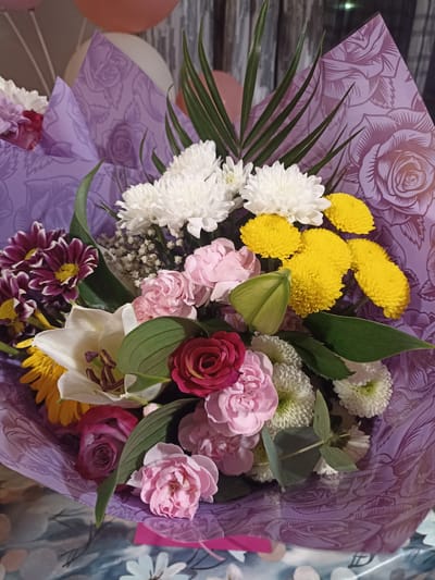 This is a medium-shot, eye-level, vertical photograph of a bouquet of flowers. The bouquet is wrapped in purple paper with a rose pattern. The flowers include white chrysanthemums, yellow button chrysanthemums, pink carnations, a white lily, a red rose, and a dark purple daisy-like flower with yellow centers. Green foliage, including fern fronds and eucalyptus leaves, is also present. In the background, out-of-focus balloons in shades of pink and orange are visible. The scene appears to be indoors, with a patterned surface in the foreground that could be a table. There are no people visible in the image. The location of Annagry, Ireland is not depicted or suggested by any elements within the photograph.