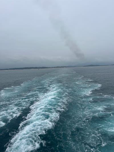The image shows a turbulent sea with a wake trailing behind a vessel, suggesting a boat or ferry is in motion. The water is a deep blue-green, with white foam and spray churned up by the movement. In the distance, a coastline is visible, with a strip of land dotted with trees and buildings. The sky is overcast and a hazy gray, with a plume of dark smoke rising in the upper left quadrant of the image, possibly from the vessel itself or a distant industrial source. The scene appears to be a coastal view in a somewhat hazy or cloudy atmosphere. There is no visible text.