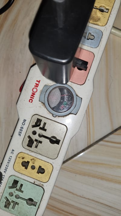 A close-up, overhead shot shows a dirty, white, multi-socket power strip with a black adapter plugged into one of its sockets. The power strip is labeled "TRONIC" and "NO: 605F" and "AC 125V/230V 13A MAX". A small analog meter is embedded in the power strip, with a needle indicating a voltage reading. The power strip is positioned on a tiled surface with a beige and brown striped pattern. The background is out of focus, but appears to be a floor with similar tiling. There are no people present in the image. The scene is indoors, likely a home or office, given the presence of the power strip and tiled floor. The location of Kipala Mpakani, Tanzania is not discernible from the image.