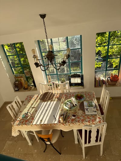 This image captures a brightly illuminated room, identified as being in Kfar Vradim, Israel. The room, likely a dining or activity area, is centered around a rectangular table with a floral tablecloth featuring patterns of red, orange, green, and cream.
On the table, there is a large white sheet of paper displaying a pencil drawing of what appears to be a vase with a plant or floral arrangement. Adjacent to the drawing are various art supplies including a cup holding paintbrushes, small containers of pigments, and an eraser. Other items on the table include a small woven placemat, a stack of books or a magazine opened to an illustrated page, a glass plate holding assorted fruits (citrus and red berries), and another glass plate with a roll of paper towel and a clear container of small items.
Surrounding the table are five white, painted wooden chairs with curved backs; one features a dark red cushion. An additional small chair with an orange seat and a black metal frame is partially visible tucked under the table on the left side.
Three distinct windows with dark green frames are set into the white walls, revealing lush green foliage outside. The leftmost window is angled inward at the top, creating a triangular section of wall above it. Strong sunlight streams through these windows, casting prominent, parallel shadows from the window frames across the table and the light-colored tiled floor.
A rustic, dark-toned metal chandelier with exposed Edison-style light bulbs hangs from the white ceiling, centered above the table.
The window sills display various objects. The left sill holds framed artwork, a wicker basket containing items, a gourd-like decorative object, and a black container with writing implements. The middle sill features a black decorative box with cut-out shapes (possibly a miniature stage or light box) and a small potted green plant. The rightmost sill is lined with numerous upright books, a wicker basket filled with textiles (an orange bandana is visible), and another small potted plant. A yellow ball is visible on the floor near the rightmost chair.