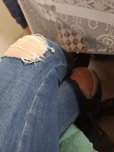 The scene is located indoors within the city of Izbeglii, Bulgaria. The primary subject is an individual's legs, bent at the knees, and clad in light blue distressed denim jeans. The left knee of the jeans features a horizontal rip with visible white frayed threads, exposing skin. The right knee also has a large, irregular rip, revealing skin. In the upper background, the underside of a surface, likely a table, is visible, covered by a tablecloth featuring a patterned design that includes grey motifs and brown-and-white checkered sections. A portion of another individual's limb, dressed in dark blue fabric, is visible in the upper left background. In the lower right, beneath the right leg, a dark object, potentially footwear, and a section of muted green fabric are visible. The posture of the legs indicates the individual is seated or reclined.