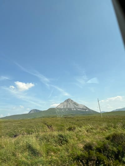 A daytime, eye-level, outdoor shot captures a panoramic view of a mountainous landscape under a bright blue sky dotted with wispy white clouds. The dominant feature is a large, grey, pointed mountain with visible lighter-colored striations, centrally located in the mid-ground. To its left, a smaller, rounded, dark grey mountain is visible. The foreground is a lush, green, undulating field of tall grass, with some areas appearing dry or brown, suggesting a natural, uncultivated environment. A single wooden telephone pole stands on the right side of the frame, angled towards the right, with thin wires extending horizontally across the scene. The overall impression is one of a rural, undeveloped, and possibly remote location. The scene is likely captured from a moving vehicle, as indicated by the slight blur in the foreground grass. The location is Meenagall, Ireland.