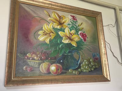 This is a framed oil painting depicting a still life. The painting features a bouquet of yellow lilies in a blue pitcher, accompanied by a basket of red cherries, two peaches, a bunch of green grapes, and a glass of red liquid. The background is a gradient of soft pinks and greens. The painting is framed in a golden-brown, ornate frame. The painting is hanging on a white wall, with a thick white pipe running vertically along the right side of the frame. A thin, dark wire is also visible, running from the top right corner of the image down the wall. The image does not provide any information about the location in Uhersko, Ukraine.