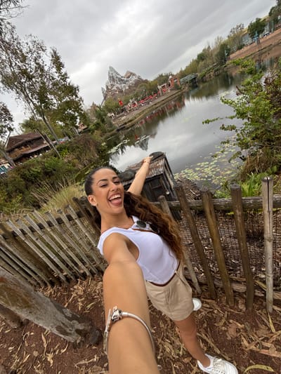 A female subject, appearing to be in her 20s or 30s with long, dark, curly hair, is captured in a selfie. She wears a white sleeveless top, light tan shorts, and white sneakers. Her left arm is extended towards the camera, revealing a silver charm bracelet on her wrist. She is smiling broadly with her mouth open and tongue slightly extended. Her right arm is raised and pointed backward towards the background.

Immediately behind her is a rustic fence constructed from vertical wooden logs and horizontal poles. The ground beneath her is reddish-brown earth with scattered leaves. To her right, behind the fence, is a small, weathered wooden structure with a rusty corrugated metal roof.

Beyond the fence and structure, a body of still water with lily pads covers the midground, reflecting the overcast sky. On the far side of the water, a prominent, artificial mountain peak, identifiable as Expedition Everest, dominates the background. Associated structures, including buildings with red roofs, lush trees, and a green metal truss bridge spanning the water, are also visible. The sky is uniformly overcast.

The scene is located at Disney's Animal Kingdom theme park in Bay Lake, United States.