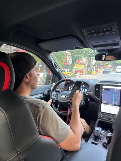 A view from inside a vehicle shows a man in the driver's seat, seemingly operating or engaging with a smartphone while stationary or moving slowly in Vị Xuyên Commune, Vietnam. He is seen from the side and slightly from behind, wearing a gray t-shirt and khaki shorts, with a black cap covering his head, leaning back against the seat's headrest. His left hand holds a smartphone displaying an application with visible text "ALERTS" and "REAR TV", while his right hand rests on the steering wheel, which features a "FORD" logo.
The car's interior is modern, with a large, vertically oriented central infotainment screen displaying a navigation map that shows a route, estimated time (e.g., "25 min"), and various app icons, including "Q.Y.O." in the top right. A digital instrument cluster behind the steering wheel shows the number "44". An overhead console has a row of six illuminated buttons labeled "AUX 1" through "AUX 6". The seats are black leather with red accents, and the gear shifter is visible on the center console.
Through the windshield, a busy street scene unfolds under bright daylight. Buildings, storefronts with indistinct signs, lush trees, and other vehicles, including a white van and a dark car, are visible, suggesting an urban or semi-urban environment. The overall impression is of a driver taking a break or navigating in traffic, using his phone.