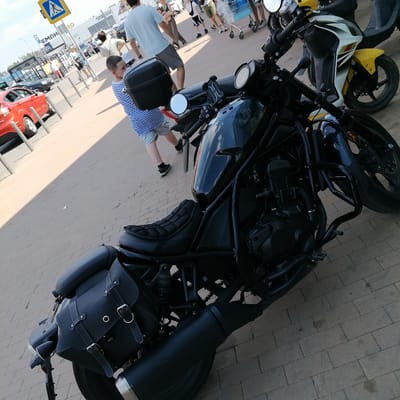 The image depicts an outdoor scene, likely in a city, with a focus on a black motorcycle in the foreground. The motorcycle has saddlebags on the side and appears to be parked on a brick-paved surface. In the background, several people are visible, some walking and others standing near what looks like a shopping cart. A red car is also parked in the background, and a pedestrian crossing sign is present. The scene suggests a public area, possibly a parking lot or street near a commercial establishment. The specific location in Yudino, Russia, is not discernible from the visual elements alone.