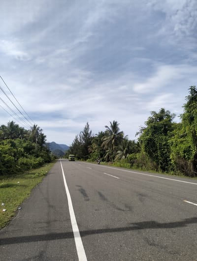 The image depicts a straight stretch of asphalt road in Meunasahmesjid, Indonesia. A solid white line divides the road. Vegetation flanks both sides of the road, transitioning into dense greenery. In the distance, a green truck and a motorbike are visible on the road. Overhanging power lines are present on the left side. The sky is overcast with patterned clouds. Mountains are subtly visible in the distance, beyond the trees. Debris is visible on the left side of the road.