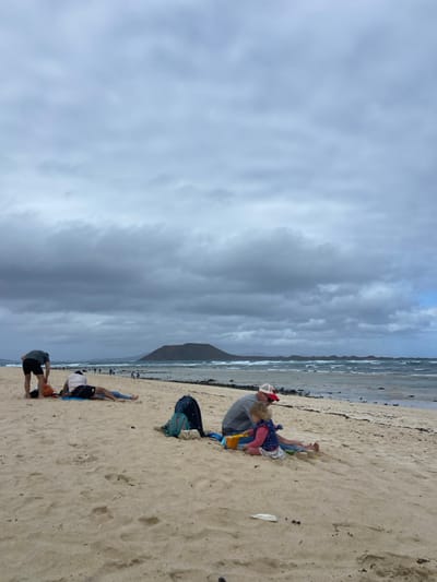 The image shows a beach scene in Corralejo, Fuerteventura, Spain (latitude: 28.724, longitude: -13.842). The sky is overcast with white clouds. The beach is a wide expanse of light-colored sand. Three people are sitting on the sand, while several more are in the distance. The person closest to the viewer is wearing a blue shirt, blue shorts, and is bending over. Next to them is another person, wearing a red shirt, black shorts, and a white hat. They are sitting on a blue and black backpack. The person furthest from the viewer is wearing a red shirt and black shorts. The scene is peaceful and tranquil.