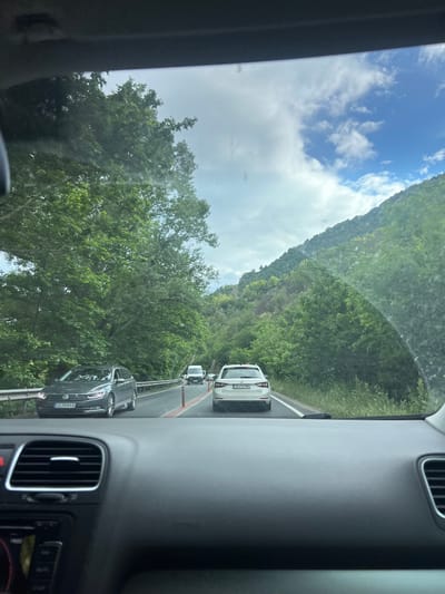 The image, taken from inside a vehicle, shows a roadway in Stara Kresna, Bulgaria. Three cars are visible in front of the camera car, with the last car closest being dark gray, the second being a white van with a visible "CB 629 MO" license plate, and the furthest a white hatchback. A green mountainside rises on the right side of the road, while trees line the opposite side, framing the partly cloudy sky. The camera is pointed slightly upwards and slightly towards the left. Parts of the dashboard and air vents of the vehicle are visible.