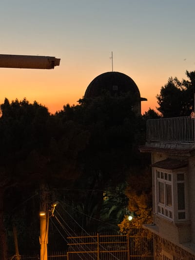 The image depicts a nocturnal scene in Izmir, Turkey, dominated by the silhouette of a domed building, possibly a observatory or similar structure, against a soft twilight sky. A tall, cylindrical chimney, with a visibly damaged top, stands prominently in the foreground, its textured surface illuminated by the fading light. The setting appears to be urban, with hints of buildings and electrical infrastructure visible. A bright streetlamp, casting a warm glow, illuminates a section of the scene, revealing foliage and a metal fence. The overall atmosphere is serene and somewhat melancholic, with the muted colors and long shadows suggesting the transition from day to night. The image seems to be taken from an elevated position, looking down upon the cityscape.