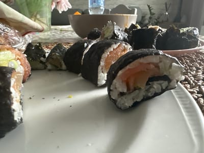 The image shows a close-up view of several pieces of sushi, likely salmon and cucumber rolls, arranged on a white plate. Some of the sushi pieces are partially eaten. In the background, there is a brown bowl, a clear plastic bottle of water, and a vase with pink flowers. The setting appears to be indoors, possibly a home kitchen or dining area. The lighting suggests it is daytime. There is no indication of the location being Macclesfield, United Kingdom.