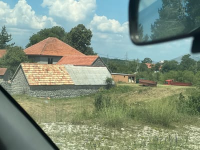 The image shows a rural scene in Vilusi, Montenegro. In the foreground, a grassy hill slopes upwards, dotted with small white and yellow wildflowers. The middle ground features a cluster of buildings with tiled roofs. One building has a stone lower wall and a tiled roof, appearing older. Adjacent to it is another building with a gray corrugated roof and a stone base. Further back, a long, narrow, light brown building with a corrugated roof is visible, and next to it are two agricultural trailers, one green and one red, which appear to be stationary. A red truck with a trailer is also parked nearby. The background is filled with lush green trees and hills, with power lines and pylons visible in the distance. The sky is blue with scattered white clouds. A reflection of trees and sky can be seen in a car's side mirror, which frames the upper right portion of the image. No people are visible in the scene.