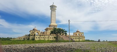 This is a photograph of the Grand Mosque of Kruengmate, located in the district of Muara Batu, Aceh Utara, Indonesia. The mosque is characterized by its tall, cream-colored minaret topped with a dome, and a sprawling, light yellow structure with multiple domes and arched windows. The building is situated in an open, grassy field with a rocky foreground. A tree with green foliage stands to the right of the main structure. In the distance, a utility pole with wires is visible, and a small vehicle, possibly a motorcycle with a sidecar or a small cart, is parked in the field. The sky is a bright blue with scattered white clouds.