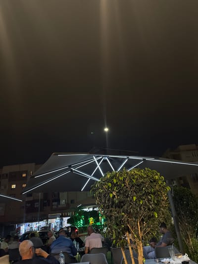 This image captures an outdoor cafe or restaurant scene at night in Saâda, Morocco, presented with a 90-degree counter-clockwise rotation, making the sky appear on the left and the ground on the right.

In the foreground, a large, modern outdoor umbrella or canopy dominates the right half of the frame. It features striking linear LED lights that illuminate its structure, casting a bright glow. Adjacent to the umbrella is a large, dense, spherical green plant or shrub, possibly in a planter, providing a natural element to the urban setting.

Several people, mostly men, are seated at tables under and around the umbrella. They appear to be socializing or relaxing. One man in a light pink shirt is visible near the center-right, while another in a blue shirt is further down. A man with a bald head and grey beard is visible towards the bottom right. Drinks, including water bottles, are visible on some tables.

The background reveals a bustling street scene. Buildings with illuminated windows line the street, and various vehicles are present. Distant streetlights and the lights from shops create a vibrant glow, with some green lights visible further down the street. The dark night sky occupies the left portion of the frame, with a bright point of light, possibly the moon or a strong light source, creating a noticeable lens flare.

The overall atmosphere suggests a lively evening, with people enjoying the outdoor ambiance. Some blurry Arabic script is faintly visible on a sign in the distant background, but it is too indistinct to be read.