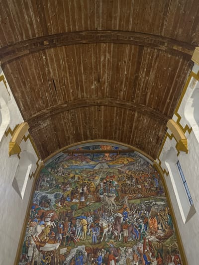 The image displays an interior view of the library within the Museo de Artes e Industrias Populares, located in Pátzcuaro, Michoacán, Mexico. The perspective is oriented upward, showcasing a prominent mural on an arched wall, surmounted by a distinctive wooden ceiling.
The ceiling is a barrel vault constructed from numerous aged, dark brown wooden planks exhibiting visible grain and areas of lighter wear. A horizontal wooden support beam crosses the arch near its apex. Flanking the mural, the walls are finished in white stucco, featuring decorative yellow architectural trim with stepped, chevron-like patterns and curved supports. A tall, narrow, multi-pane window is visible on the right wall.
The central feature is a large, colorful mural, identified as Juan O'Gorman's "The History of Michoacán." It spans the entire arched wall, depicting a complex historical narrative. The upper section portrays a landscape with blue water bodies, green terrain, and a fiery sky or volcanic eruption in the background. Indigenous figures are engaged in activities such as hunting, with a jaguar visible in the upper left.
The mid-section of the mural is dominated by scenes of the Spanish Conquest. Armored Spanish conquistadors on horseback, wielding swords and lances, are depicted in conflict with indigenous Purépecha warriors. The indigenous figures wear feathered headdresses and carry round shields and spears. Acts of violence are illustrated, including falling figures and a scene in the lower right showing individuals on a pyre, accompanied by a skeletal figure symbolizing death.
The lower section of the mural features various figures, including laborers, individuals in traditional dress, and a figure holding a book, likely representing historical or intellectual figures. A banner inscribed with "UTOPIA" is also present. The mural employs a rich color palette of greens, blues, reds, browns, and ochres.
The scene is illuminated by a combination of interior lighting and natural light from the window on the right, casting subtle shadows on the adjacent white wall.