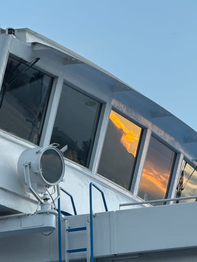 This close-up image captures a section of the white superstructure of a vessel, likely a ship or large boat, set against a clear, light blue sky. The photograph was taken in the location context of Brek, Russia.
The main subjects are the ship's upper deck, featuring multiple rectangular windows and structural elements. The top two visible windows display a prominent, vibrant golden-orange reflection, strongly suggesting either a sunrise or sunset, as if reflecting brightly lit clouds or a low sun from the opposite direction. This warm light contrasts with the darker, more muted reflections in the lower windows.
A large, circular white spotlight or searchlight, mounted on an arm, is a prominent object in the lower-middle section of the frame. To the right, sections of blue metal railings are visible, providing a pop of color against the otherwise white and grey tones of the ship. A sturdy, vertical white column or mast structure is also present on the right side, with some smaller structural details and pipes. Windshield wipers are discernible on the top-most window, indicating a bridge or pilot house area. The ship's exterior is painted in a uniform light grey or white, with minor visible wear. The weather appears clear and bright. There are no people visible, and no discernible text. The scene is static, showing no specific activity.