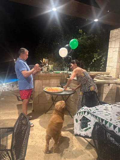 Observed outdoors at night, likely a backyard in Southlake, United States of America, the scene features a man, a woman, and a dog. The man, in a blue shirt and red shorts, appears to be taking a photograph with a mobile phone. A woman in a floral dress is serving food from a platter on a table. A medium-sized, tan-colored dog is nearby on the ground. Green, black, and white balloons are visible, along with a table with a graduation-themed tablecloth. The background includes stone structures and foliage, illuminated by artificial lighting.