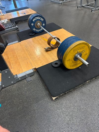 The image depicts a weightlifting setup in a gym. A barbell is loaded with weight plates. On one side are blue plates labeled "ROGUE 45 LB," and on the other side are yellow plates labeled "HAMMER STRENGTH 55." There are additional smaller plates near the barbell. A weightlifting belt is also visible. The equipment is situated on a platform with a wooden section and a black rubber mat. The gym has gray flooring and other gym equipment in the background. The location appears to be inside a weightlifting facility in the city of Duluth, United States of America.