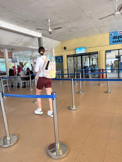 This media file depicts a scene inside a ferry terminal, likely in Tambon Don Sak, Thailand, given the visible text in Thai and English. The main subject is a young woman from behind, wearing a white long-sleeved shirt, maroon shorts, and white sneakers. She has a backpack with a cartoonish pattern over her shoulders and her hair is pulled back with a scrunchie. She is standing in a queue, marked by silver stanchions and blue rope barriers.
Several other people are visible in the background, some also appearing to be waiting in line or seated. One person is wearing a red shirt, and others are in casual attire. The setting is indoors, with a tiled floor and a yellow wall with signs. A ceiling fan is visible, suggesting a warm climate.
The prominent text indicates "SAMUI," suggesting the destination of the ferry. There's also a sign for "GATE 1" with boarding and gate closing times: "BOARDING 17:00" and "GATE CLOSE 16:55." This implies the time of day is late afternoon. The overall atmosphere seems to be one of organized travel, with passengers waiting for their ferry departure. The lighting suggests it is daytime.