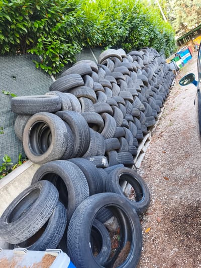 A large pile of used tires is stacked along a chain-link fence, bordered by a green hedge. The tires are of various sizes and brands, some appearing newer than others. They are arranged in a somewhat organized but dense manner, extending in a long line.

In the background, beyond the tires, there are some structures that appear to be part of an industrial or service area. One structure has colorful panels, and a sign with blue and white text that may read "ESOTAM". There is also a blue car visible, parked on the right side of the image, with its side mirror reflecting the surroundings.

The ground in front of the tires is covered with gravel and small stones, with some fallen leaves and pine needles scattered around. The lighting suggests it is daytime, likely afternoon, with sunlight casting shadows. The overall scene appears to be an outdoor storage area for discarded tires, possibly a recycling or repair facility. The location is identified as Chiugiana, Italy. There is no visible text on the tires themselves that is legible in this image. No people are present.