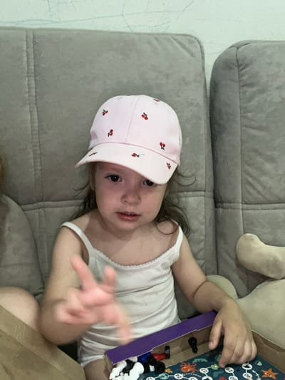 A young child, wearing a pink hat with cherry patterns and a white tank top, lies on a grey couch. The child's face is turned towards the camera, with dark eyes and slightly parted lips. Their right hand is outstretched, appearing blurred as if in motion. Beside the child, a board game with numbers and sea creatures is visible, along with some game pieces. A soft toy can be seen in the background on the couch. The scene appears to be indoors, possibly in a living room, given the couch and the presence of a board game. The lighting suggests it is daytime. The overall mood is casual and playful. There is no visible text in the image.