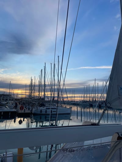 The image depicts a marina in La Grande-Motte, France, observed during twilight conditions, indicated by a low sun on the horizon. The viewpoint is from the deck of a vessel, with its white boom, rigging lines, and a section of a greyish-white sail, featuring a blue emblem, visible in the foreground and extending upwards on the right side. The marina is densely populated with numerous sailboats, uniformly docked in the calm, reflective water. Their masts and rigging form a prominent series of vertical and diagonal lines across the middle ground and background. The sky transitions from a clear blue overhead to areas of scattered grey and purple clouds, with a distinct band of orange and yellow light visible at the horizon line. The still water accurately mirrors the boats, masts, and sky above. In the mid-ground, a white-hulled sailboat is clearly identifiable, displaying a red circular lifebuoy attached to its stern. No individuals are visible within the scene, and all vessels appear stationary.