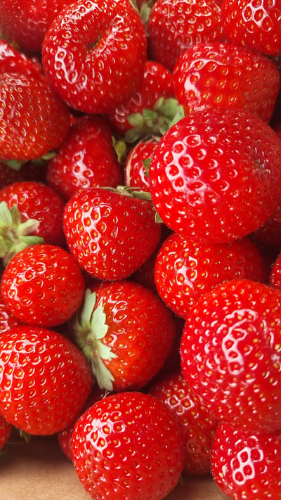 A close-up, top-down view of a large pile of ripe, red strawberries. The strawberries are varied in size and shape, with some appearing larger and rounder, while others are more elongated. The surface of each strawberry is covered in numerous small, yellow seeds. The green calyxes, or leafy tops, of some of the strawberries are visible, adding a touch of contrasting color to the predominantly red scene. The lighting highlights the glossy texture of the strawberries, creating bright reflections on their surfaces. The background appears to be a neutral, light brown surface, possibly a box or tray, which further emphasizes the vibrant color of the fruit. There are no people or any discernible urban elements; the image is focused solely on the strawberries, suggesting it was taken in a setting where the fruit is being prepared or displayed, such as a market stall or a kitchen. The image is consistent with an agricultural context and could be from any location globally.