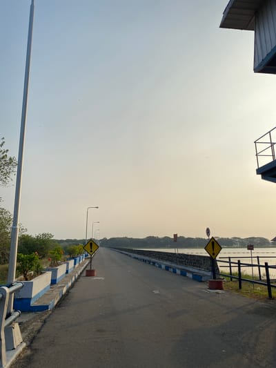 The image depicts a long, paved road that appears to be a causeway or bridge, bordered by a railing on the right and a raised concrete barrier with planters on the left. The causeway stretches into the distance, leading towards a tree-lined horizon. Several streetlights are visible along the left side of the road, and two yellow diamond-shaped warning signs with black exclamation points are placed at intervals on the causeway. In the background, a body of water is visible to the right of the causeway, with trees and a structure with a metal roof visible in the far distance on the right. The sky is clear and light, suggesting it is either early morning or late afternoon. The overall scene is serene and tranquil. The location is identified as the Bendungan (dam) in Bandungan, Indonesia.