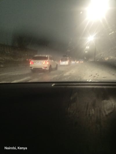 The image presents a nighttime road scene, viewed from inside a vehicle looking forward through a rain-streaked windshield. The bottom third of the frame is occupied by the dark dashboard or interior part of the car.
On the wet road ahead, several vehicles are visible. A white sedan, possibly a Subaru, with illuminated red taillights, is prominent in the mid-ground. Behind it, a white van or SUV can be seen, followed by the fainter lights of other cars further down the road. The road surface appears highly reflective due to the wet conditions.
The scene depicts traffic moving slowly or stopped during a rainstorm at night. Heavy rain is evident from the numerous water droplets and streaks on the windshield, which distort the view. Bright streetlights on the right side of the road cast intense glares, creating starburst effects and significantly reducing overall visibility. These artificial lights brightly illuminate sections of the wet road and contribute to the hazy atmosphere.
Notable details include the challenging weather conditions (heavy rain), the time of day (night), and the low visibility caused by the combination of darkness, rain, and bright glare from streetlights. The overall impression is one of cautious travel in adverse weather. Visible text in the bottom left corner of the original image reads "Nairobi, Kenya." The provided location context for this description is Benoni, South Africa.